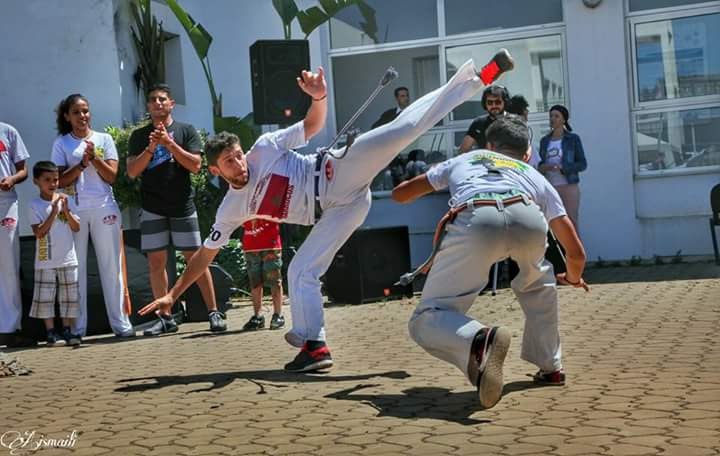 Ayoub Chatar en plein jeu de capoeira, jambe tendue au-dessus de son partenaire, devant un public