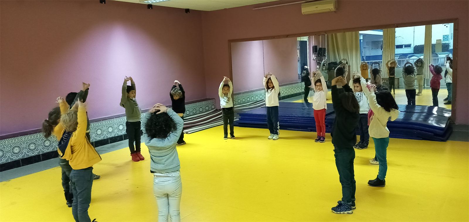 Élèves de maternelle en cercle, bras levés au-dessus de la tête, pendant l'échauffement dans une salle de motricité