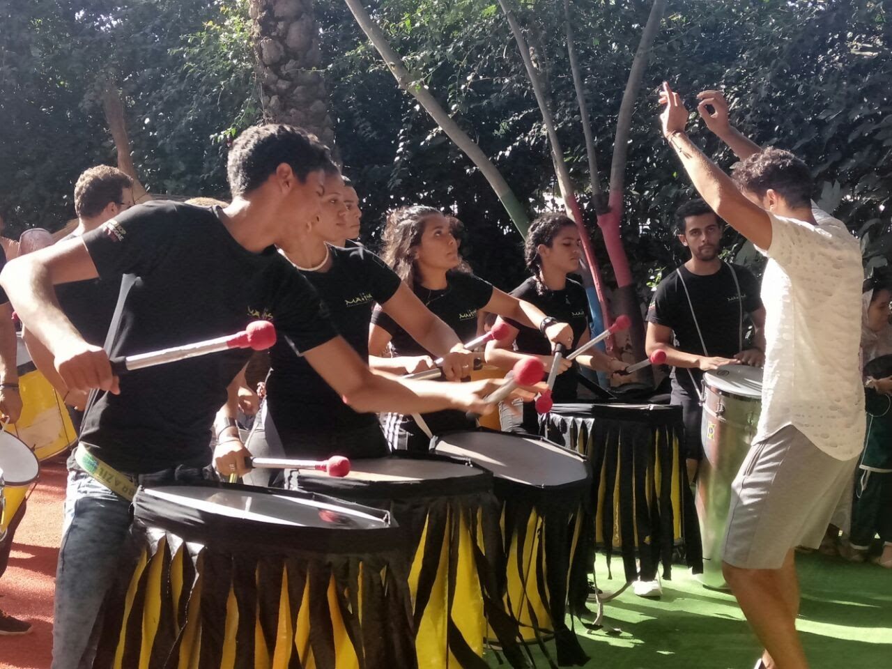 Un groupe d'adultes frappe ensemble sur des tambours, baguettes levées, pendant un atelier de percussions en plein air