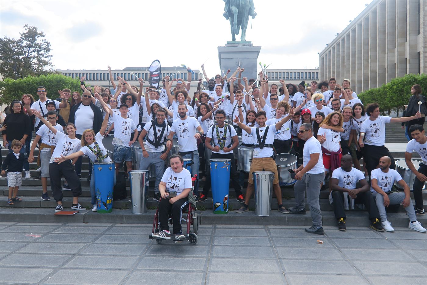 Un grand groupe de participants en t-shirts blancs pose, bras levés, derrière ses percussions sur une place