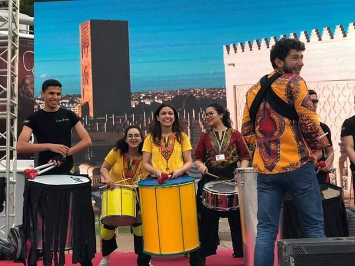 Ayoub Chatar, en chemise wax, mène la troupe de percussionnistes sur scène avec la tour Hassan de Rabat en arrière-plan